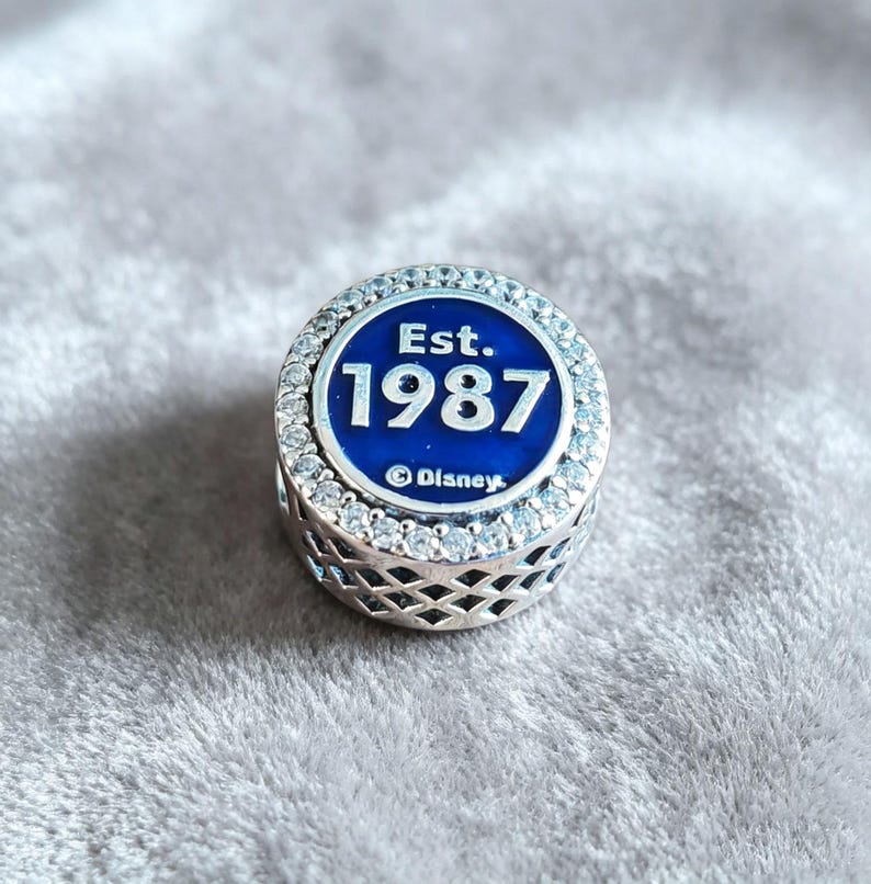 Pode incluir: Um berloque de prata com um centro de esmalte azul com o texto "Est. 1987" e o logotipo da Disney. O berloque &eacute; circundado por um anel de pedras claras e brilhantes e tem um design decorativo vazado nas laterais.