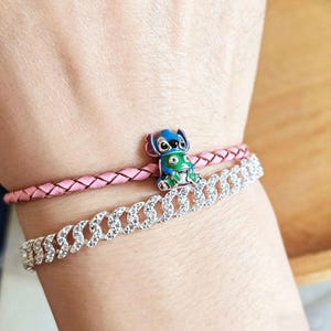 May include: Two bracelets are shown. The top bracelet is pink braided leather. The bottom bracelet is silver with clear stones. A small charm of a blue and green cartoon character is on the pink bracelet.
