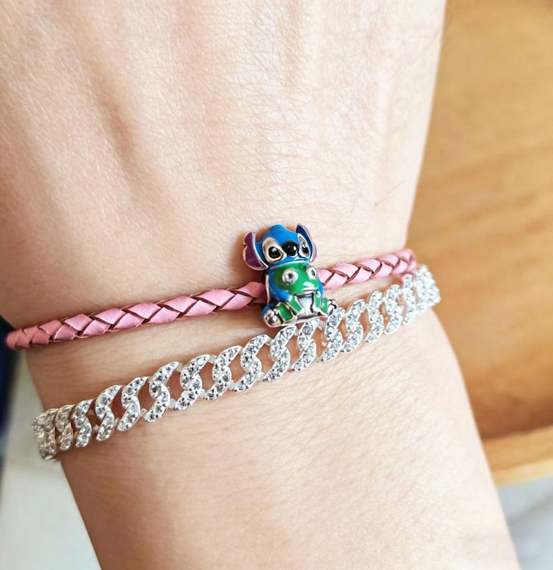 May include: Two bracelets are shown. The top bracelet is pink braided leather. The bottom bracelet is silver with clear stones. A small charm of a blue and green cartoon character is on the pink bracelet.