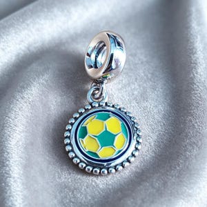 Puede incluir: Un charm de plata con un diseño de balón de fútbol. El charm presenta un colgante redondo con un diseño de balón de fútbol verde y amarillo, rodeado por un borde plateado con cuentas. El charm tiene un lazo plateado en la parte superior para sujetarlo.