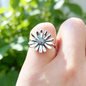 May include: A silver ring featuring a daisy flower design with elongated petals radiating from a textured, circular center. The metal has a polished finish with darkened accents in the crevices of the petals and the center to highlight the floral details.