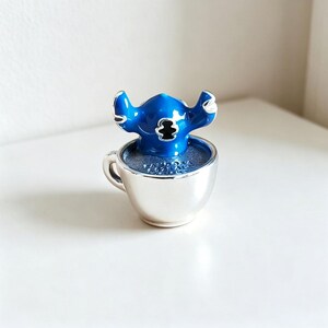 Pode incluir: Uma figura azul de Stitch est&aacute; dentro de uma x&iacute;cara de ch&aacute; prateada. A figura tem orelhas grandes e um emblema preto no peito. A x&iacute;cara de ch&aacute; &eacute; preenchida com um l&iacute;quido azul e est&aacute; sobre uma superf&iacute;cie branca. &Eacute; um item colecion&aacute;vel.