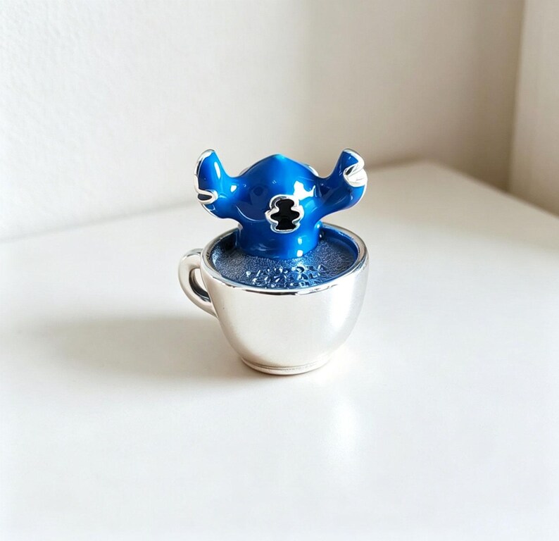 Pode incluir: Uma figura azul de Stitch est&aacute; dentro de uma x&iacute;cara de ch&aacute; prateada. A figura tem orelhas grandes e um emblema preto no peito. A x&iacute;cara de ch&aacute; &eacute; preenchida com um l&iacute;quido azul e est&aacute; sobre uma superf&iacute;cie branca. &Eacute; um item colecion&aacute;vel.