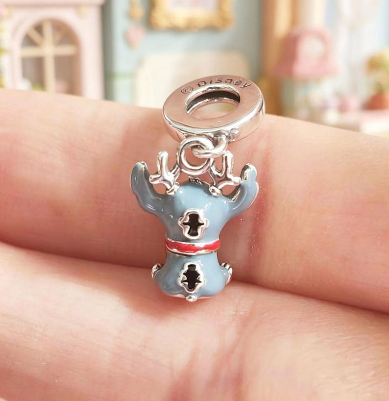 K&ouml;nnte beinhalten: Ein silberner Charm mit einem blau-roten Emaille-Stitch-Design. Der Charm hat oben eine kleine Schlaufe mit dem Disney-Logo. Die Stitch-Figur hat einen roten Kragen und hebt die Arme.