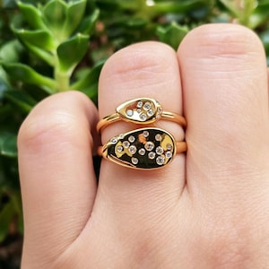 May include: Two gold rings featuring teardrop-shaped settings. The smaller top ring has a cluster of five small clear stones, while the larger bottom ring features a wider teardrop shape with a scattered arrangement of twelve small clear stones.