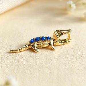 Pode incluir: Um berloque de metal em tom dourado com o formato de um pequeno jacaré ou crocodilo. A criatura apresenta uma série de detalhes em esmalte azul brilhante ao longo das costas e uma pequena argola na cabeça para fixação. A superfície é polida.