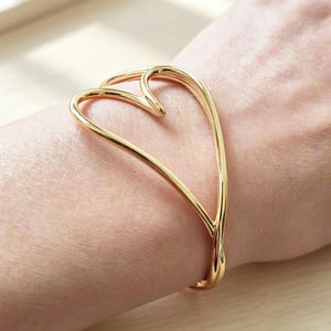 Puede incluir: Un brazalete rígido de tono dorado pulido con un diseño de alambre abierto continuo que forma una gran figura de corazón estilizada. La banda de metal es lisa y redondeada, envolviendo la muñeca con una estructura abierta.