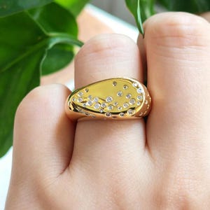 Puede incluir: Un anillo de tono dorado con una forma orgánica irregular que presenta un grupo de pequeñas piedras claras de corte redondo incrustadas en la superficie. El metal tiene un acabado liso y pulido, con las piedras dispuestas en un patrón denso.