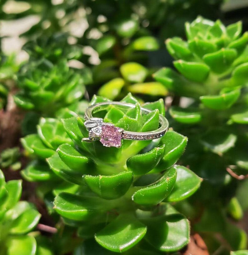 Puede incluir: Un anillo de plata con una gema rosa cuadrada y peque&ntilde;as piedras transparentes. El anillo presenta un peque&ntilde;o dise&ntilde;o de conejito en un lado. El anillo est&aacute; sobre un fondo de plantas suculentas verdes.