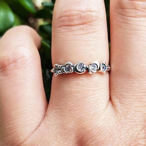 Puede incluir: Un anillo de tono plateado que presenta una serie de formas de media luna alternadas y pequeñas piedras transparentes de corte redondo engastadas a lo largo de la banda. El metal tiene un acabado pulido y un patrón de motivos celestiales.