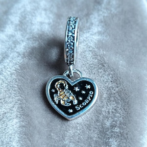Op de afbeelding: Een zilveren hartvormige bedel met een zwarte emaille inleg, een goudkleurige schorpioenfiguur, kleine sterren en het woord SCORPIO in zilver gegraveerd. Het oogje is versierd met een rij kleine, heldere, fonkelende steentjes.