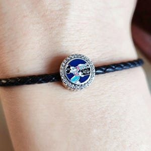 Pode incluir: Uma pulseira de couro tran&ccedil;ado preta com um berloque redondo. O berloque tem um fundo azul e um design do Mickey Mouse com uma borda prateada e cristais transparentes. O berloque tem a palavra "Disney".