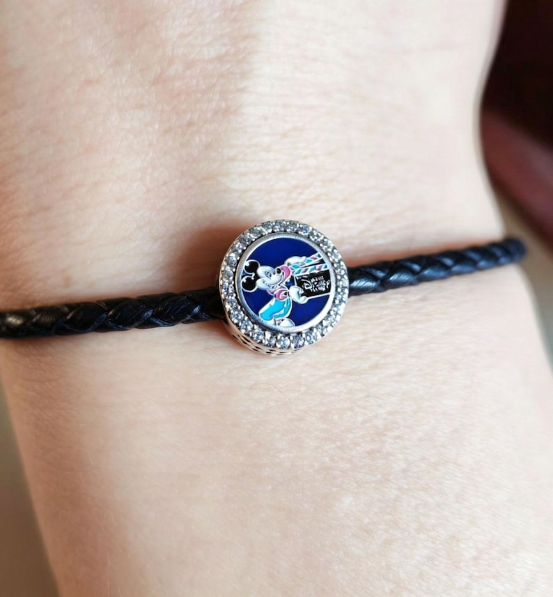 Pode incluir: Uma pulseira de couro tran&ccedil;ado preta com um berloque redondo. O berloque tem um fundo azul e um design do Mickey Mouse com uma borda prateada e cristais transparentes. O berloque tem a palavra "Disney".
