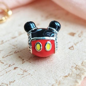 May include: A silver charm bead shaped like a stylised Mickey Mouse head with black enamel ears, a red enamel face, and two yellow oval eyes. The side of the silver metal frame is engraved with the text PANDORA and Disney in black lettering.