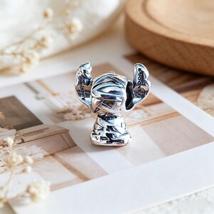 May include: A silver charm depicting a stylized character with large ears and a textured body. The charm is marked with the Disney and Pandora logos. The charm is on a white and beige background.