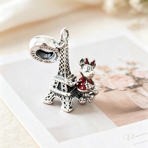 May include: A silver charm featuring the Eiffel Tower and a Minnie Mouse figure in a red jacket. The charm has a loop for attaching to a bracelet or necklace. The word "Disney" is visible on the charm.
