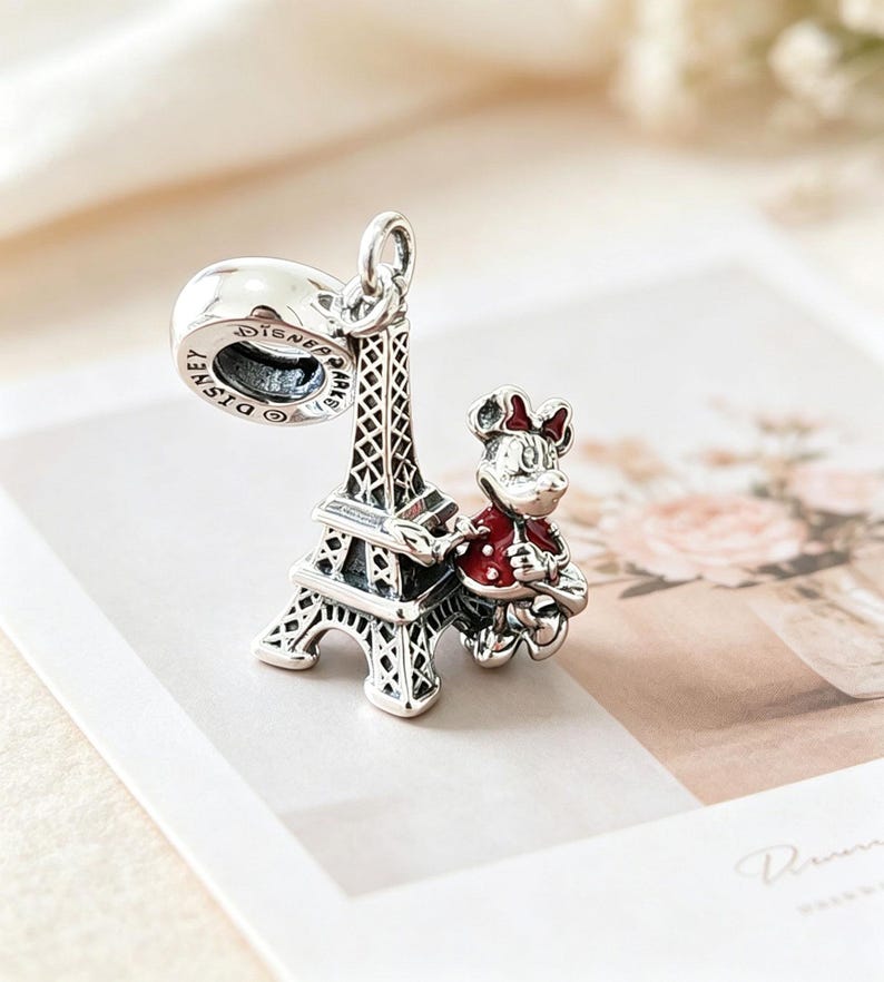May include: A silver charm featuring the Eiffel Tower and a Minnie Mouse figure in a red jacket. The charm has a loop for attaching to a bracelet or necklace. The word "Disney" is visible on the charm.