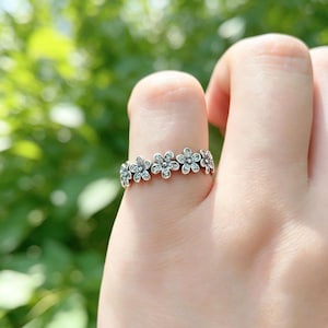Puede incluir: Un anillo de tono plateado que presenta una banda continua de pequeñas formas de flores de cinco pétalos. Cada flor individual está acentuada con una pequeña piedra transparente y brillante en su centro, creando un patrón floral delicado.