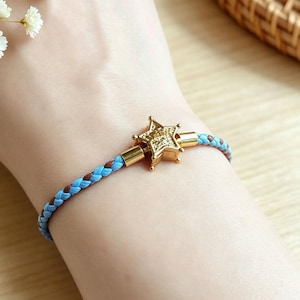 Puede incluir: Una pulsera trenzada con un dije central de estrella en tono dorado con superficie texturizada. La banda está construida con cordones entrelazados de color azul claro y marrón, asegurados por terminales metálicos dorados unidos a la estrella.