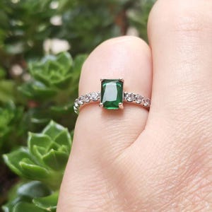 May include: A gold-coloured ring with a rectangular, emerald-green gemstone as the focal point. The band is embellished with small, clear gemstones. The ring is set against a blurred green backdrop.