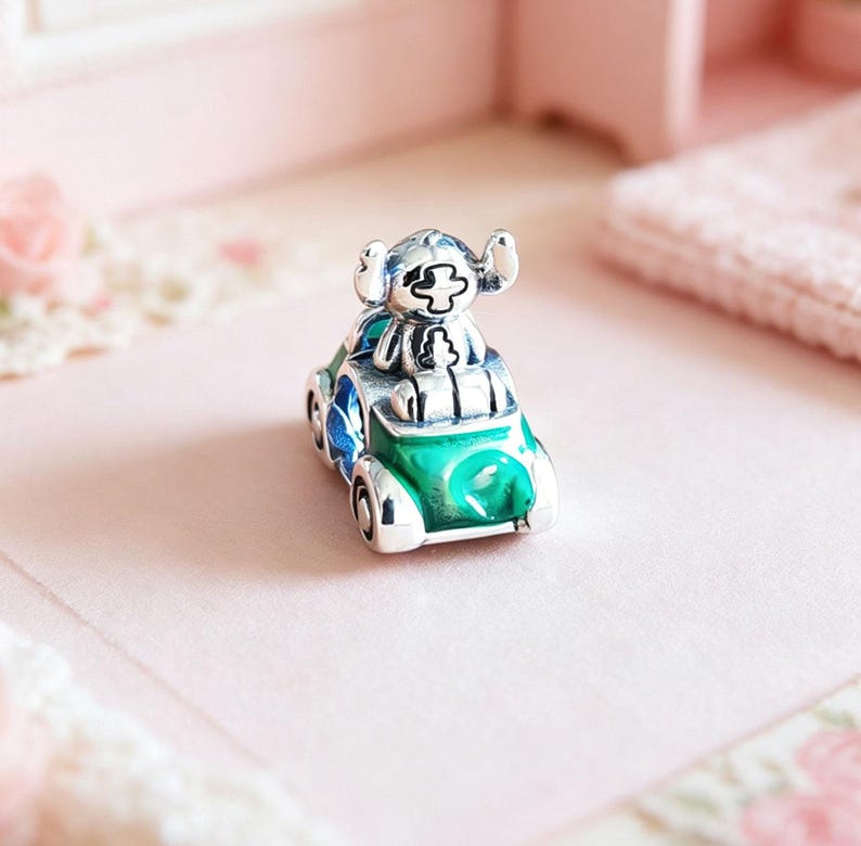 May include: A silver charm featuring a cartoon character driving a green car with blue accents. The charm has a detailed design with enamel coloring and is likely a collectible item or jewelry accessory.