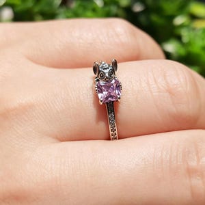 Puede incluir: Un anillo plateado con una peque&ntilde;a figura de animal y una gema cuadrada de color p&uacute;rpura claro. La banda del anillo est&aacute; adornada con peque&ntilde;as piedras transparentes. El anillo se muestra en un dedo.