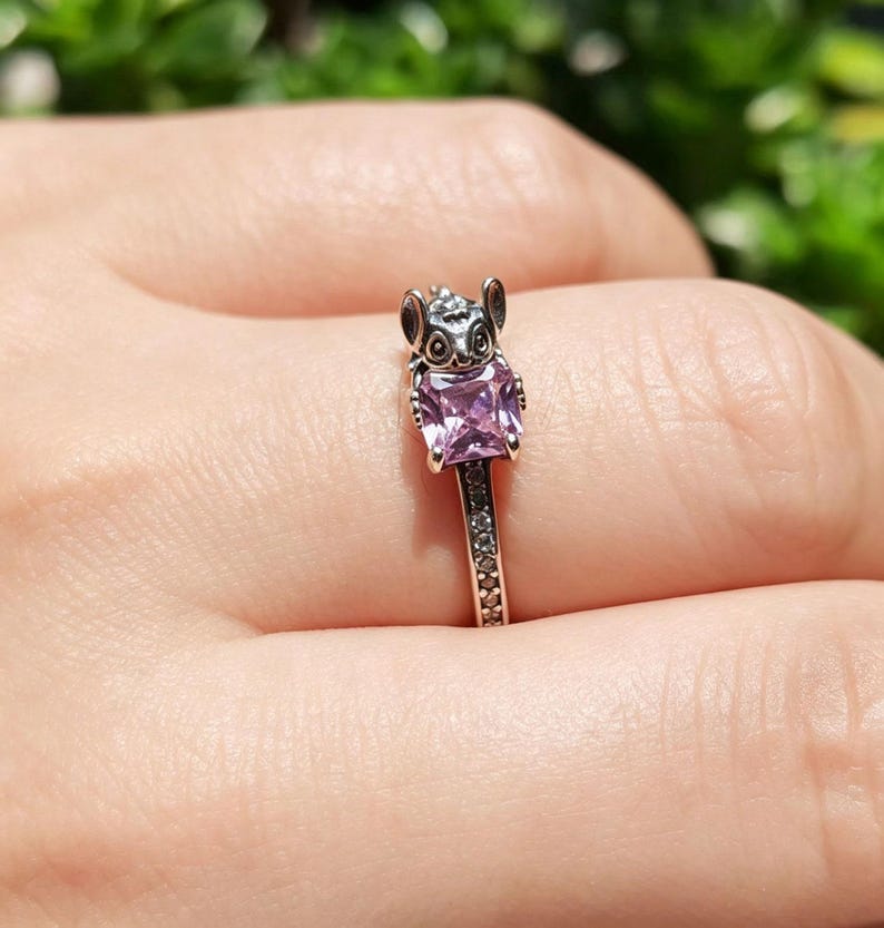 Puede incluir: Un anillo plateado con una peque&ntilde;a figura de animal y una gema cuadrada de color p&uacute;rpura claro. La banda del anillo est&aacute; adornada con peque&ntilde;as piedras transparentes. El anillo se muestra en un dedo.