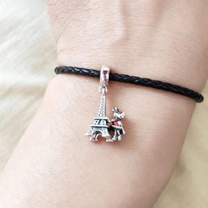 May include: A silver charm with the Eiffel Tower and a small dog with a red bow tie. The charm hangs from a black braided leather bracelet. A small red heart is at the top of the charm.