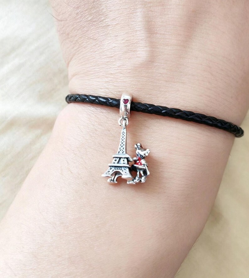 May include: A silver charm with the Eiffel Tower and a small dog with a red bow tie. The charm hangs from a black braided leather bracelet. A small red heart is at the top of the charm.
