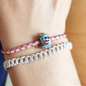 Puede incluir: Una mu&ntilde;eca adornada con tres pulseras. Una pulsera de cuero trenzado rosa, una pulsera de cadena plateada con piedras transparentes y un charm azul con un personaje de dibujos animados con sombrero y bufanda roja.