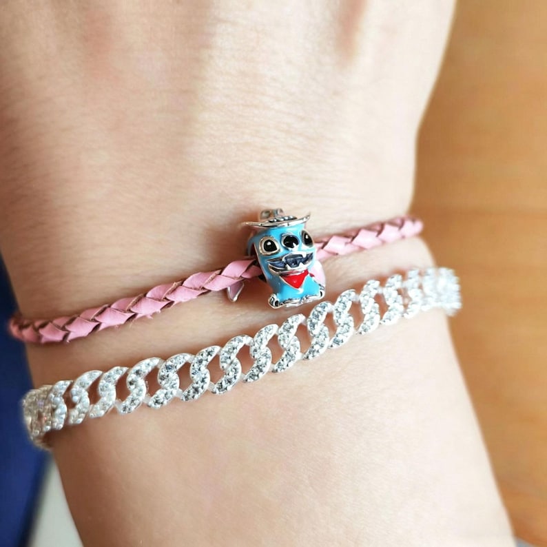 Puede incluir: Una mu&ntilde;eca adornada con tres pulseras. Una pulsera de cuero trenzado rosa, una pulsera de cadena plateada con piedras transparentes y un charm azul con un personaje de dibujos animados con sombrero y bufanda roja.