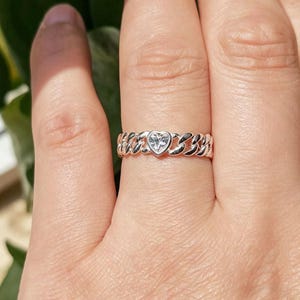 May include: A silver ring with a heart-shaped gemstone at the center. The ring features a chain-link design around the band. The ring is on a finger.