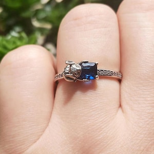 May include: A silver ring featuring a small bunny design on one side and a square, deep blue gemstone on the other. The band is adorned with small, clear stones. The ring is shown on a finger.