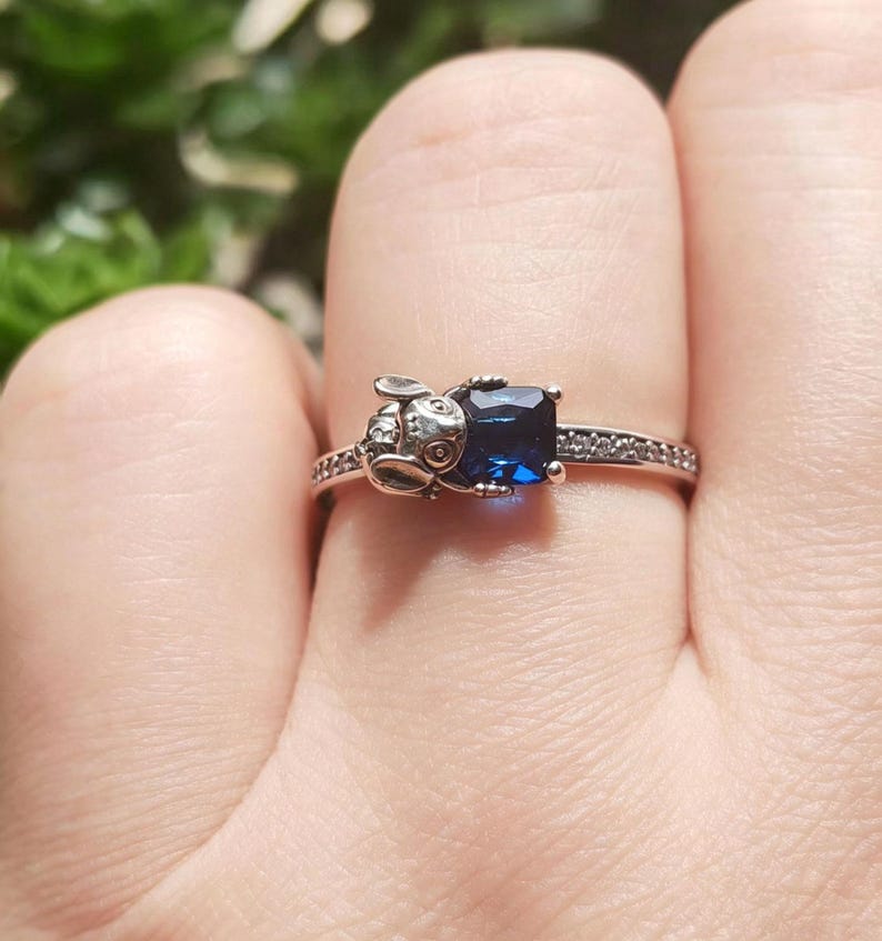 May include: A silver ring featuring a small bunny design on one side and a square, deep blue gemstone on the other. The band is adorned with small, clear stones. The ring is shown on a finger.