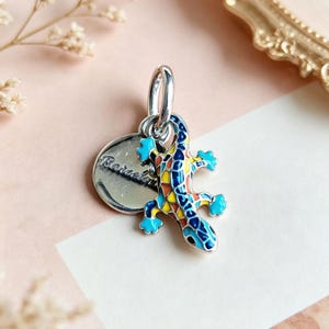 Könnte beinhalten: Ein silberner Anhänger in Form einer Eidechse mit blauem, gelbem und orangefarbenem Emaille-Mosaikmuster. An der Eidechse ist eine kleine, runde, polierte Silberscheibe mit der Gravur Barcelona befestigt. Der Anhänger hat einen runden Ring.