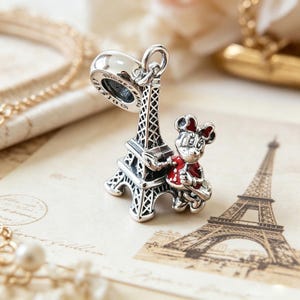 May include: A silver charm featuring the Eiffel Tower and a cartoon character in a red outfit. The charm has a loop for attaching to a bracelet or necklace. The background includes a printed image of the Eiffel Tower.