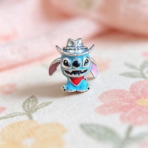Puede incluir: Un charm de personaje de dibujos animados azul con un sombrero de vaquero, un pa&ntilde;uelo rojo y orejas rosas. El charm tiene una expresi&oacute;n sonriente y est&aacute; hecho de metal plateado y esmalte.