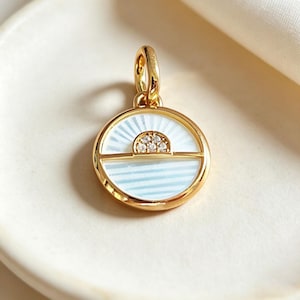 Può includere: Un ciondolo circolare in oro con un inserto in smalto bianco e una linea orizzontale che divide il cerchio. La sezione superiore presenta un motivo a raggiera, mentre quella inferiore ha strisce orizzontali. Un piccolo semicerchio dorato con cinque pietre chiare incastonate si trova al centro.