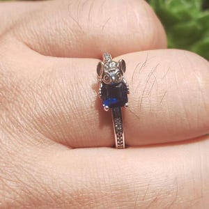 May include: Silver ring featuring a koala bear design with a dark blue rectangular gemstone. The ring band is adorned with small, clear gemstones. The koala has detailed features, including ears and eyes.