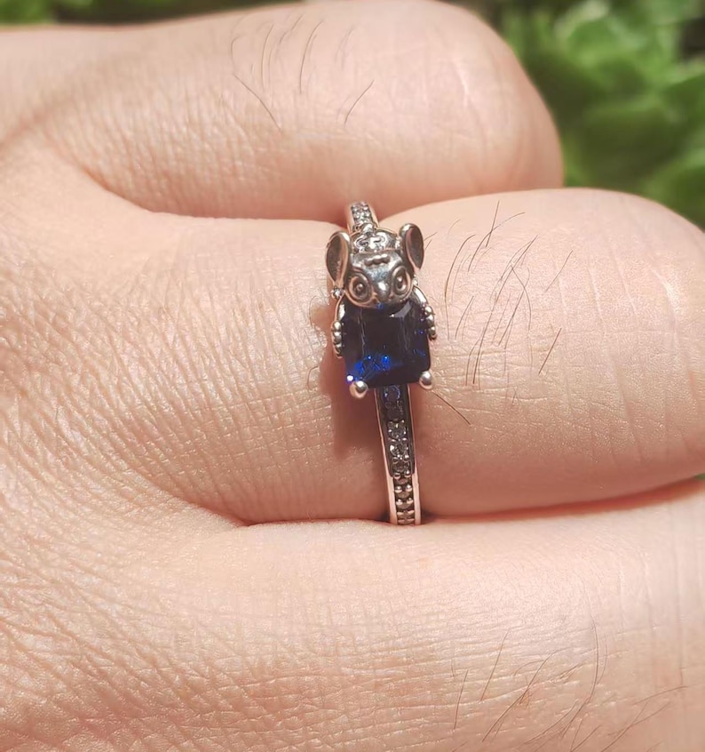 May include: Silver ring featuring a koala bear design with a dark blue rectangular gemstone. The ring band is adorned with small, clear gemstones. The koala has detailed features, including ears and eyes.