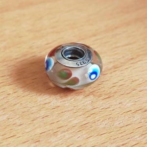 Op de afbeelding: Een heldere glazen kraal met een zilverkleurige metalen kern. De kraal heeft kleurrijke accenten van rood, blauw en groen. De metalen kern is gestempeld met "S925". De kraal is waarschijnlijk voor een armband of ketting.