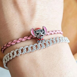 Op de afbeelding: Twee armbanden om een pols. De bovenste armband is een roze gevlochten leren band met een Stitch-bedel. De onderste armband is een zilveren ketting met heldere strass-steentjes.