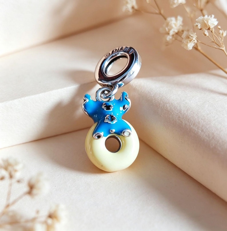 May include: A silver charm with a blue and yellow enamel design. The charm features a blue figure on top of a yellow donut shape. The charm has a silver loop with the word "Disney" engraved on it.