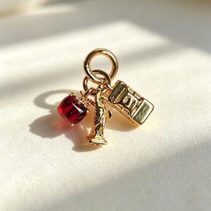 Op de afbeelding: Een goudkleurige bedelhangertje met drie bungelende elementen aan een ring: een dieprode gefacetteerde appel, een miniatuurfiguur van het Vrijheidsbeeld en een kleine gele taxi, allemaal vervaardigd uit goudkleurig metaal.