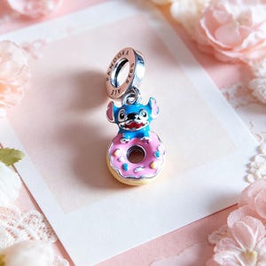 May include: A silver charm with a blue and pink enamel Stitch character sitting on a pink donut with sprinkles. The charm has a ring with the word "PANDORA" and "S925 ALE" engraved on it.