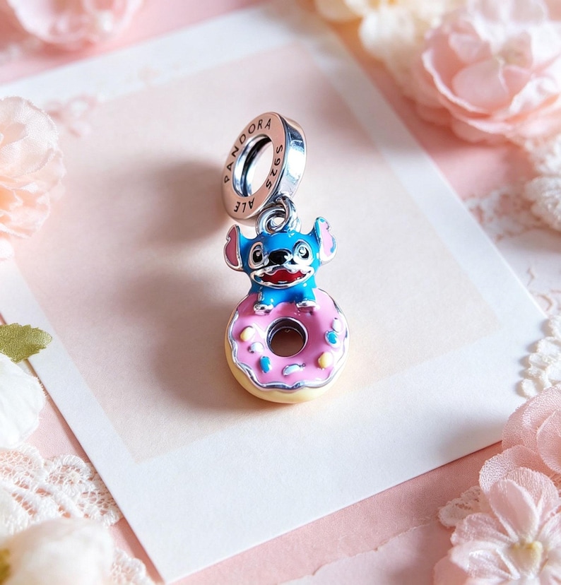 May include: A silver charm with a blue and pink enamel Stitch character sitting on a pink donut with sprinkles. The charm has a ring with the word "PANDORA" and "S925 ALE" engraved on it.