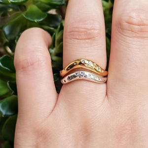 Peut inclure: Deux bagues en forme de chevron empilées, l'une en or poli et l'autre en argent poli. Chaque bague présente un design de bande ondulé et organique avec plusieurs petites pierres rondes et claires incrustées dans la surface métallique.