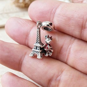 May include: A silver charm featuring the Eiffel Tower and a Minnie Mouse figure in a red dress. The charm has a heart-shaped detail and is designed to be added to a bracelet or necklace. The charm is detailed and has a polished finish.