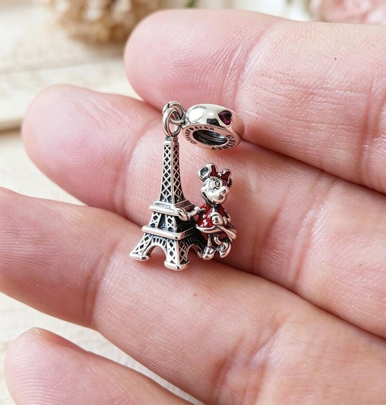 May include: A silver charm featuring the Eiffel Tower and a Minnie Mouse figure in a red dress. The charm has a heart-shaped detail and is designed to be added to a bracelet or necklace. The charm is detailed and has a polished finish.
