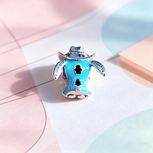 Puede incluir: Un charm de color azul claro con un personaje de dibujos animados con un sombrero de vaquero. El charm tiene detalles negros y la palabra "Disney" est&aacute; inscrita en un lado. El charm es de metal plateado, probablemente una joya.